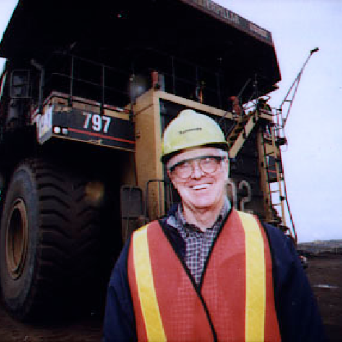 A photo of Charles Koch wearing a high-visibility orange safety vest in front of a large piece of heavy machinery at an industrial site.