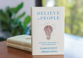 A hardcover copy of Believe in People, co-authored by Charles Koch and Brian Hooks, standing upright against a neutral gradient background.