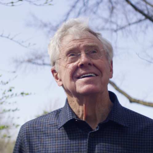 An outdoor photo of Charles Koch with white hair and glasses wearing a dark checkered shirt looking toward the sky.
