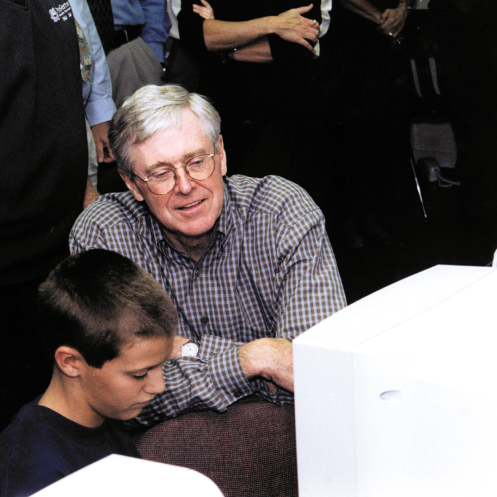 An intrigued Charles Koch looks over the shoulder of a young student working at a computer in the foreground.