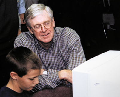 An intrigued Charles Koch looks over the shoulder of a young student working at a computer in the foreground.